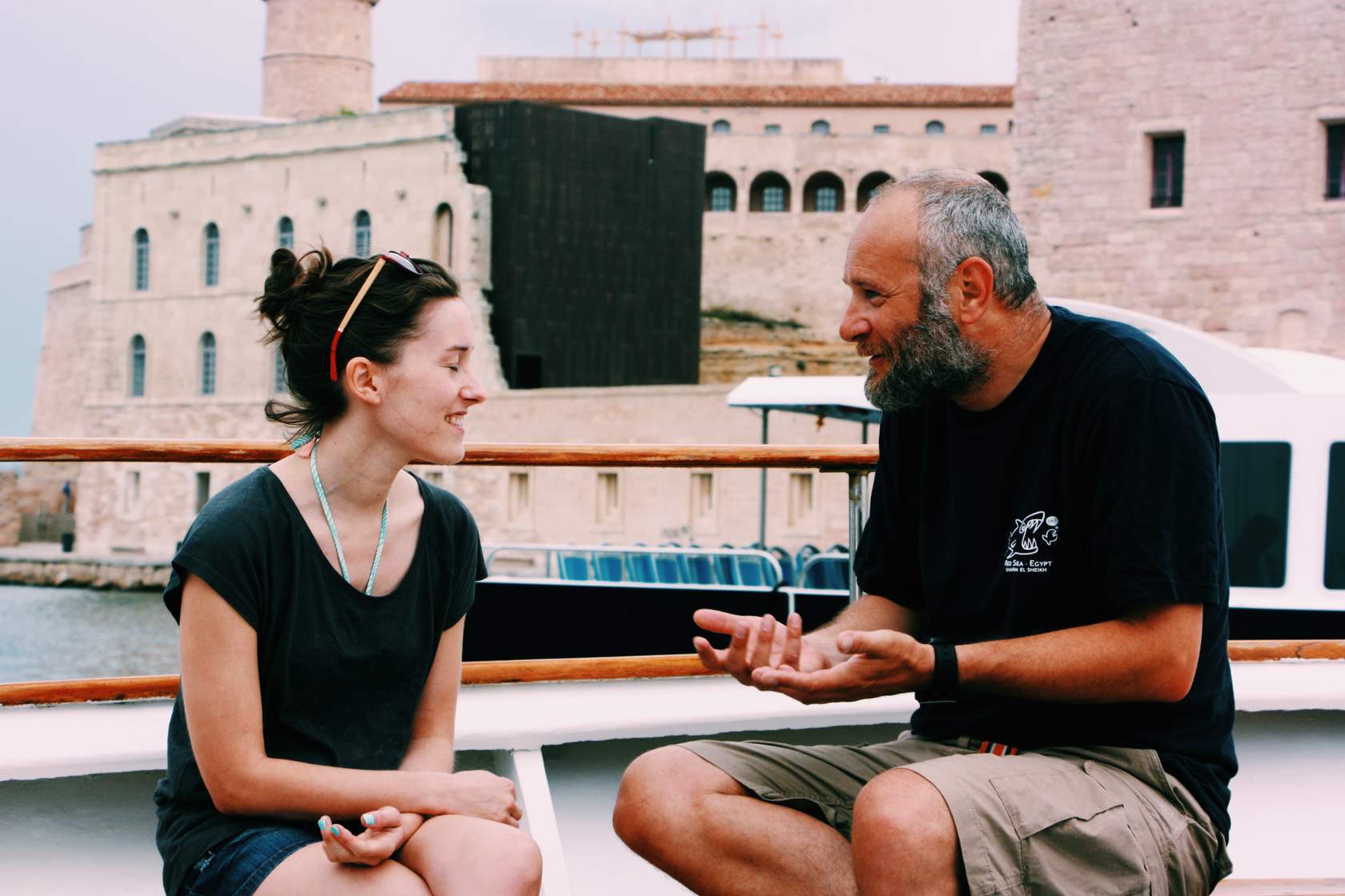 A man and woman engaged in conversation on a boat, with a castle in the background.