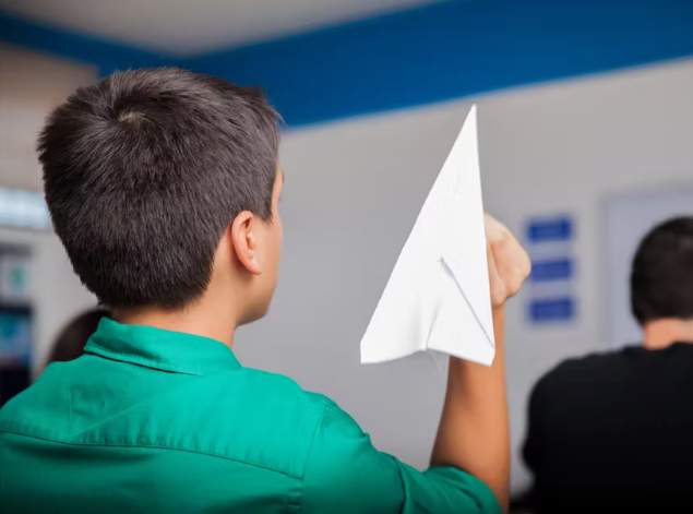 Mrs. Howell observing a boy holding up a paper airplane during consultation in front of a classroom for Part 4.
